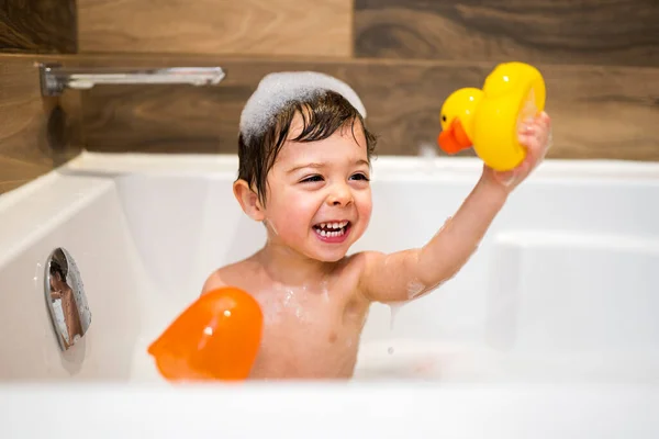 Happy two year kid bathes with toys in bath with foam and duck, — Photo