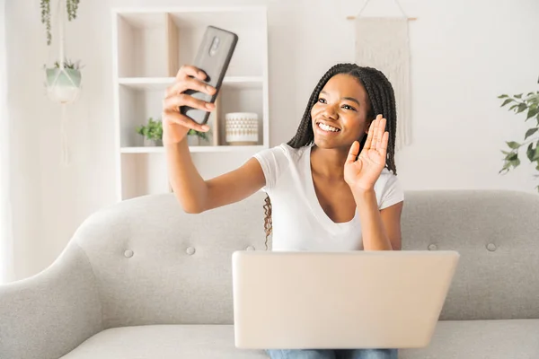 Jeune afro-américaine adolescent sur un canapé dans son salon en utilisant un ordinateur portable et un téléphone portable — Photo