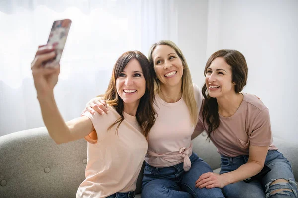 Tres alegres feliz mujer usando el teléfono inteligente móvil en casa juntos sentados en el sofá tomar una foto — Foto de Stock