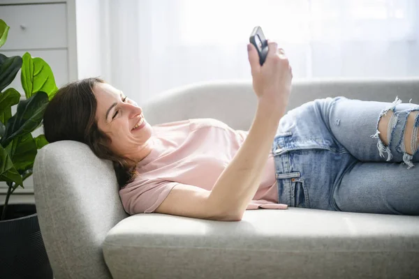 Friendly looking long-haired girl using cellphone sitting at home on couch — ストック写真