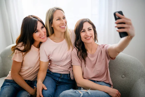Tres alegres feliz mujer usando el teléfono inteligente móvil en casa juntos sentados en el sofá tomar una foto — Foto de Stock