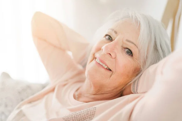 Inomhusporträtt av attraktiv glad 70-årig äldre kvinna med behagligt leende avkopplande på grå soffa i sin moderna lägenhet — Stockfoto