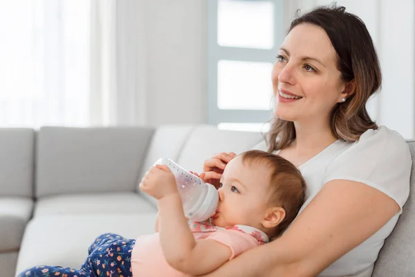 Joyeux sourire mère et bébé stationnant sur le canapé à la maison et le bébé mange du lait — Photo