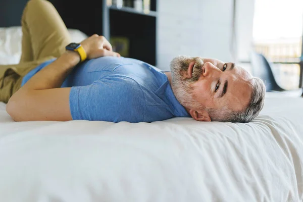Симпатичный взрослый мужчина дома с седой бородой, расслабляющийся на кровати — стоковое фото