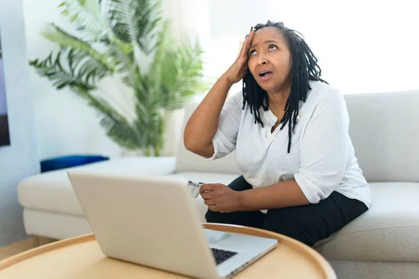 Fénykép csinos fekete nő használja laptop otthon kanapé, és vesz valamit a weben úgy néz ki, mint aggódni emiatt — Stock Fotó