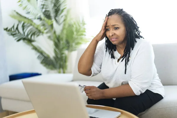 Fénykép csinos fekete nő használja laptop otthon kanapé, és vesz valamit a weben úgy néz ki, mint aggódni emiatt — Stock Fotó