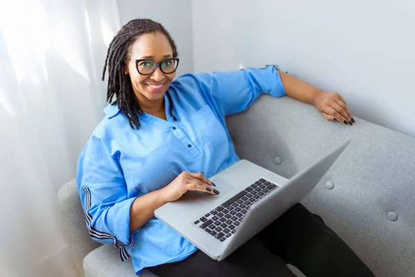 Φωτογραφία της όμορφης μαύρης γυναίκας που χρησιμοποιεί φορητό υπολογιστή στον καναπέ στο σπίτι — Φωτογραφία Αρχείου
