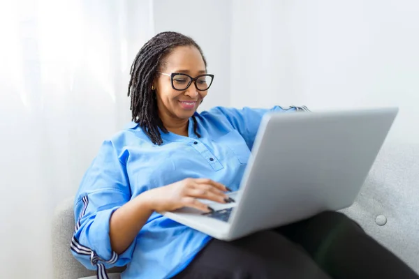 Φωτογραφία της όμορφης μαύρης γυναίκας που χρησιμοποιεί φορητό υπολογιστή στον καναπέ στο σπίτι — Φωτογραφία Αρχείου