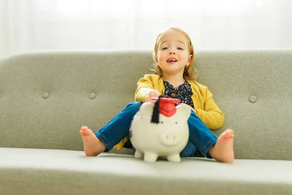 Cute little baby girl putting a coin into a pink piggybank seated on a gray sofa — Photo