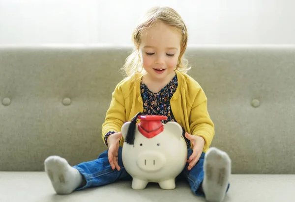 Cute little baby girl putting a coin into a pink piggybank seated on a gray sofa — Stock fotografie