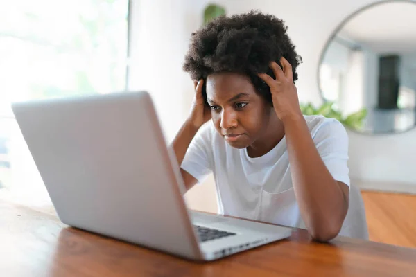 Νεαρή Αφρικανή επιχειρηματίας που εργάζονται σε απευθείας σύνδεση με ένα φορητό υπολογιστή, ενώ κάθεται στο τραπέζι της κουζίνας στο σπίτι φαίνονται λυπημένος — Φωτογραφία Αρχείου