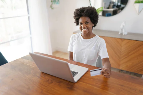 Souriante jeune entrepreneure africaine travaillant en ligne avec un ordinateur portable tout en étant assise à sa table de cuisine à la maison — Photo