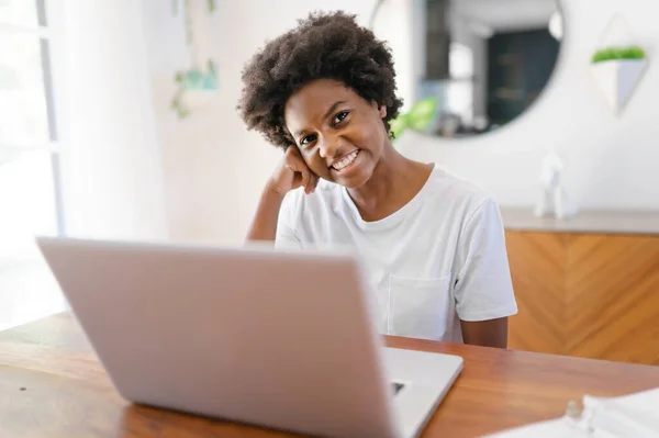 Souriante jeune entrepreneure africaine travaillant en ligne avec un ordinateur portable tout en étant assise à sa table de cuisine à la maison — Photo