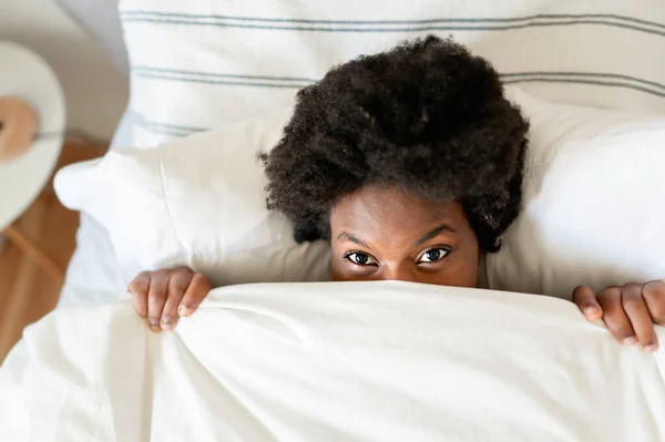 Beautiful young African woman in tank top lying in bed cover face with sheet — Stockfoto