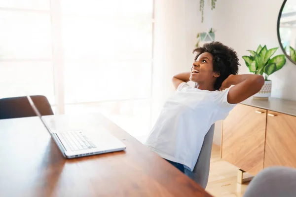 Souriante jeune entrepreneure africaine travaillant en ligne avec un ordinateur portable tout en étant assise à sa table de cuisine à la maison — Photo