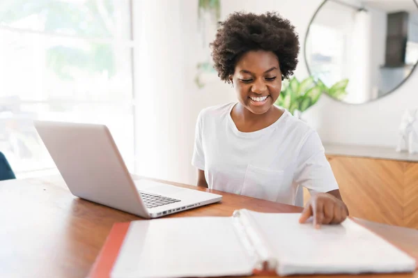 Χαμογελώντας νεαρή Αφρικανή επιχειρηματίας που εργάζεται σε απευθείας σύνδεση με ένα φορητό υπολογιστή, ενώ κάθεται στο τραπέζι της κουζίνας στο σπίτι — Φωτογραφία Αρχείου