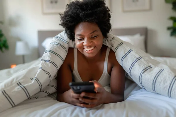 Unga afrikanska kvinna liggande på sängen med mobiltelefon — Stockfoto