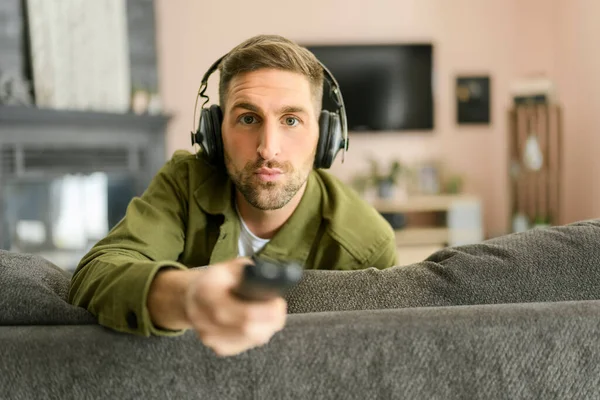 Handsome guy wearing headphones enjoys music on the comfortable sofa with remote control — Zdjęcie stockowe