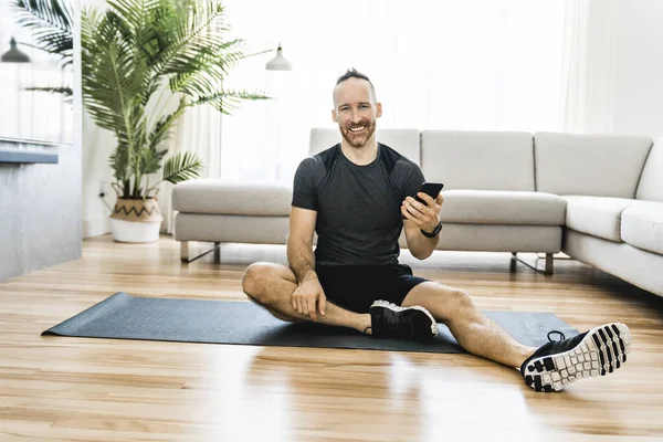 Чоловік на килимку виконує деякі вправи вдома, використовуючи мобільний телефон, щоб допомогти тренуватися — стокове фото