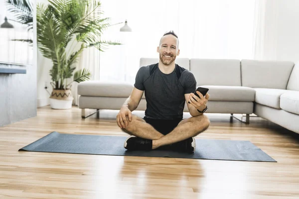 Чоловік на килимку виконує деякі вправи вдома, використовуючи мобільний телефон, щоб допомогти тренуватися — стокове фото
