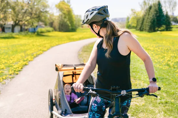 Woman riding a bicycle with a baby stroller attached to the bicycle. — Stock Photo, Image