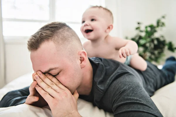 Tired father sleep but his baby boy interferes him. Young happy father lying on bed while his son play with him — Zdjęcie stockowe