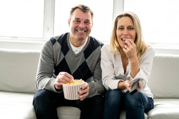 A Happy young couple lying on the sofa at home with popcorn watching TV — Stok fotoğraf