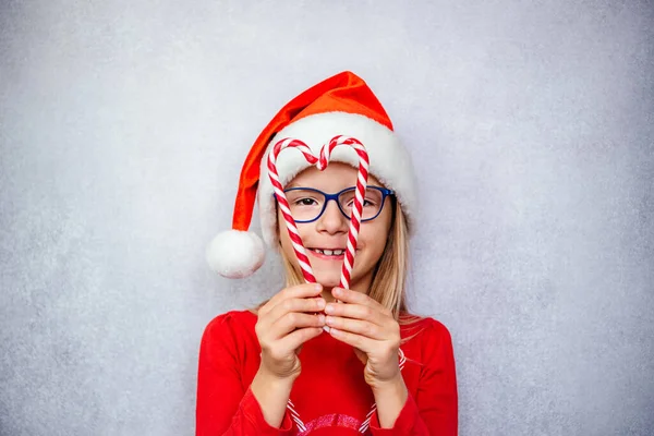 Щаслива маленька дівчинка в окулярах і капелюсі Санта робить форму серця з цукерками, Різдвяна дитина Офтальмологія — стокове фото