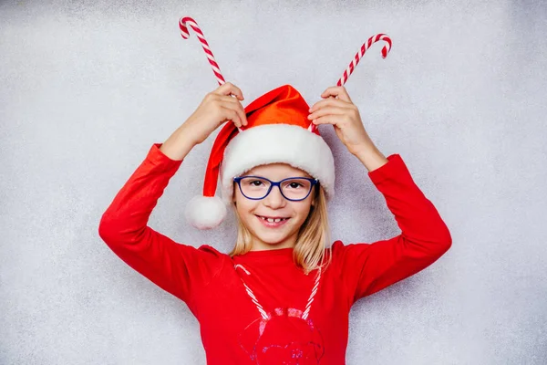 Щаслива маленька дівчинка в окулярах і капелюсі Санта і тримає цукерки на голові, щоб зробити роги для оленів — стокове фото
