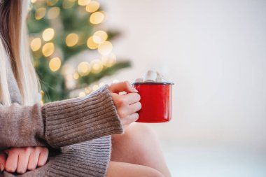Noel ağacı ışıklarında marshmallowlu kırmızı bir fincan sıcak çikolata tutan eller arka planda.