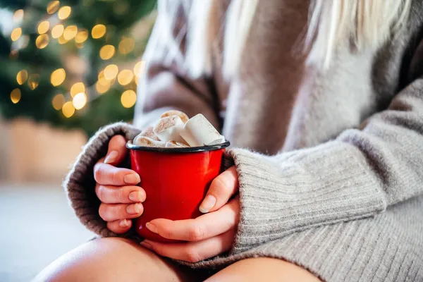 A blonde girl holding a red cup of hot chocolate with marshmallows on Christmas tree lights bokeh background — Stock Photo, Image