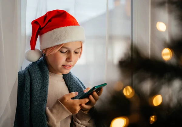 Kırmızı Noel Baba şapkalı genç kız ön planda Noel ağacı olan cep telefonu kullanıyor.