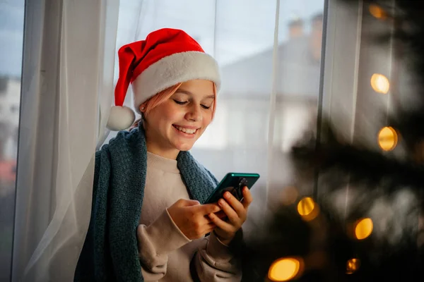 Молода щаслива дівчина-підліток в червоному капелюсі Санта використовує мобільний телефон з ялинкою на передньому плані — стокове фото