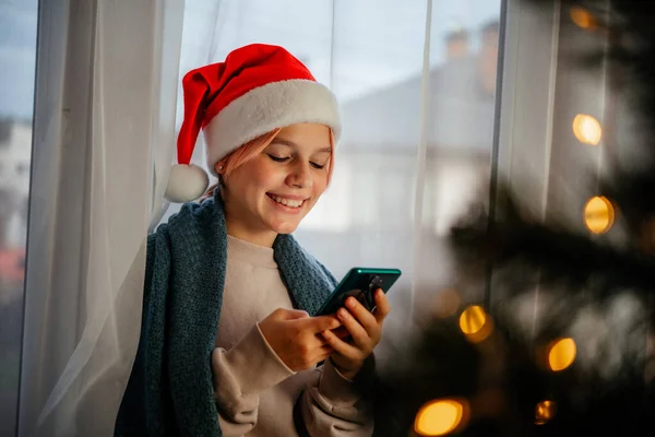 Kırmızı Noel Baba şapkalı genç kız ön planda Noel ağacı olan cep telefonu kullanıyor.