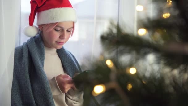 Joven adolescente feliz chica en rojo sombrero de santa usando el teléfono móvil con el árbol de Navidad en primer plano — Vídeo de stock