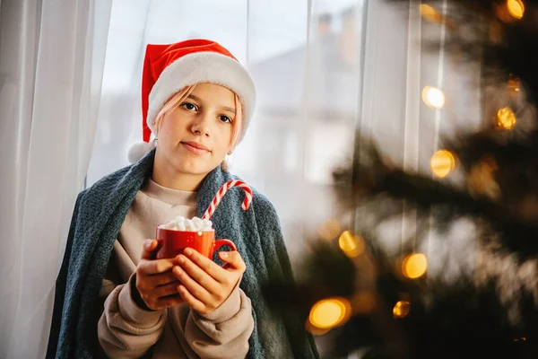 Молода дівчина-підліток у капелюсі Санта має чашку гарячого шоколаду з зефіром з ялинкою на передньому плані — стокове фото