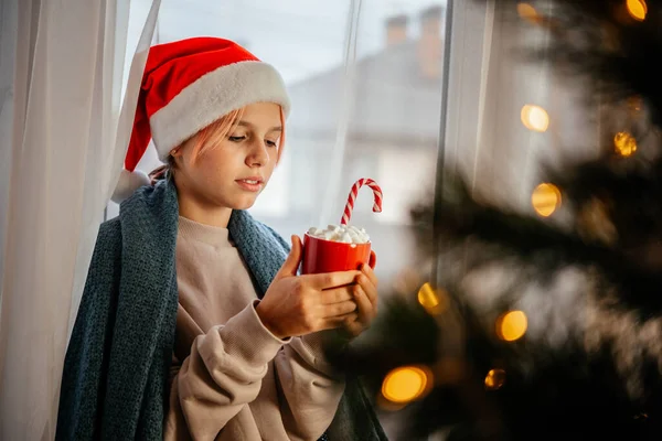 Молода дівчина-підліток у капелюсі Санта має чашку гарячого шоколаду з зефіром з ялинкою на передньому плані — стокове фото