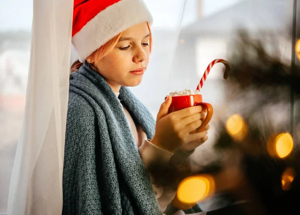 Junges Teenie-Mädchen mit Weihnachtsmütze mit einer Tasse heißer Schokolade mit Marshmallow und Weihnachtsbaum im Vordergrund — Stockfoto