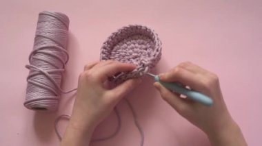 Pembe bir arka planda örgü kancası ve pembe renkli iplik tutan kadın eller, örgü ve tığ işi malzemeleri.