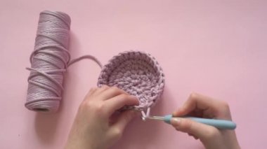 Pembe bir arka planda örgü kancası ve pembe renkli iplik tutan kadın eller, örgü ve tığ işi malzemeleri.