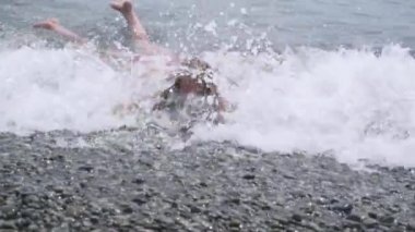 Pebbe sahilinde yatan bir çocuk köpüklü deniz dalgalarıyla arkasındaki çakıl taşlı sahile çarpıyor.