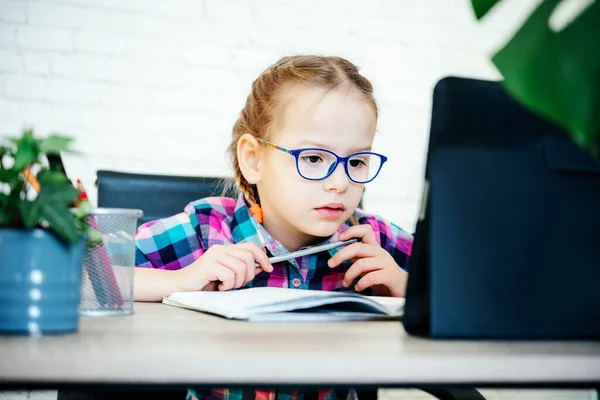 Basisschoolmeisje in een bril aan tafel met behulp van haar tablet tijdens hevige afsluiting, afstandsonderwijs voor kinderen — Stockfoto