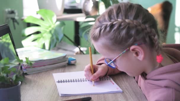 Niña de la escuela primaria en anteojos escribiendo en su cuaderno mientras tiene una lección en línea durante el bloqueo covid — Vídeo de stock
