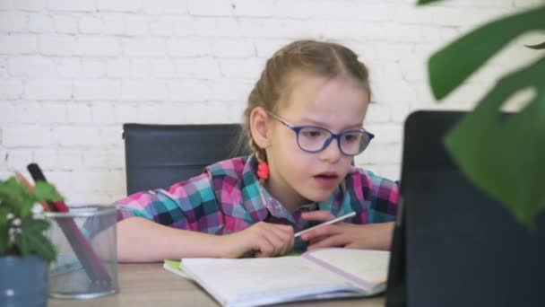 Chica de la escuela en gafas de vista mirando su tableta y tomando notas en el cuaderno mientras que tiene lección en línea — Vídeos de Stock