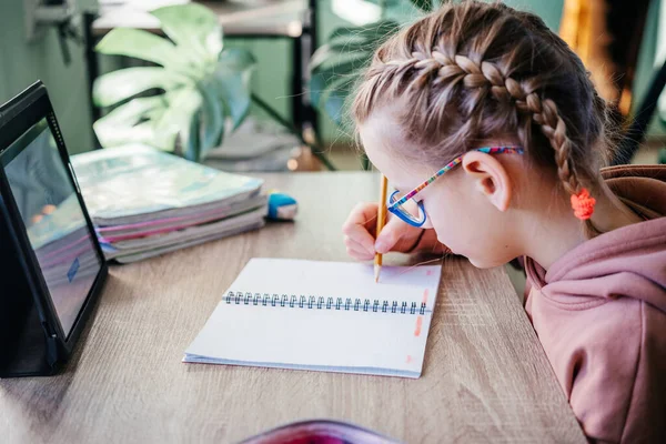 Basisschool meisje in bril schrijven in haar notebook tijdens het hebben van online les tijdens covid lockdown, afstandsonderwijs voor kinderen — Stockfoto