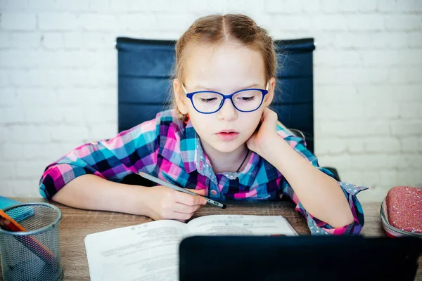 Menina em óculos estudando on-line em casa, uma menina da escola primária com computador tendo zoom vídeo chat com professor Fotos De Bancos De Imagens