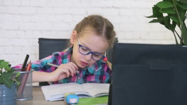 Ragazza della scuola in occhiali a guardare il suo tablet e prendere appunti nel notebook pur avendo lezione online — Video Stock