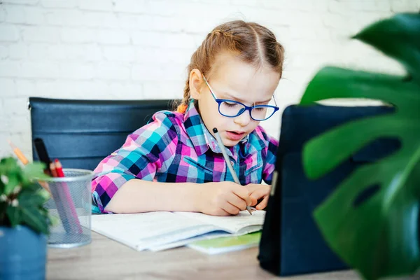 Klein meisje in bril het maken van notities in haar noteblook whlile studeren online thuis — Stockfoto