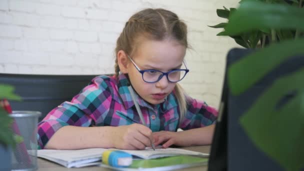 Általános iskolás lány szemüvegben ír a jegyzetfüzetébe, miközben online leckéje van a tömérdek zárlat alatt, távoktatás gyerekeknek — Stock videók