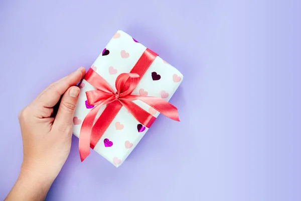 Una caja de regalo de mano envuelta en papel patrón corazón con lazo de cinta roja sobre fondo púrpura —  Fotos de Stock